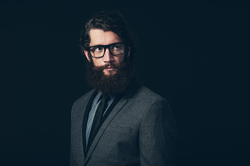 Close up Gorgeous Young Man with Long Goatee Beard, Wearing Formal Fashion with Eyeglasses, Looking to the Upper Right of the Frame on a Black Background.
