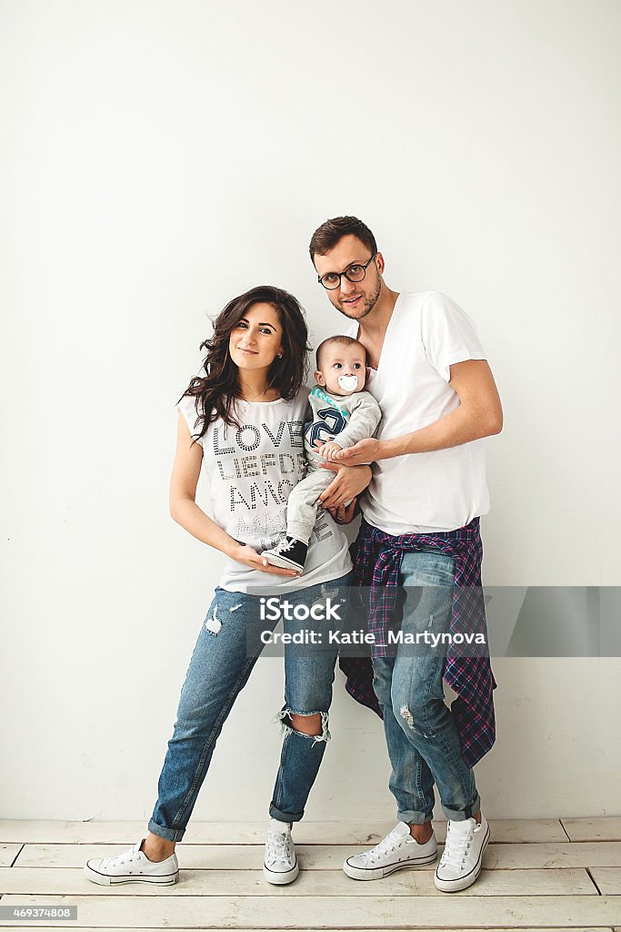 Hipster father, mother holding cute baby boy over white backgrou Young hipster father, mother holding cute baby boy on rustic wooden floor over white background Young Family Stock Photo