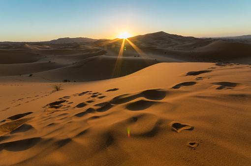 Sunrise and sunshine ray over sand dunes in Sahara Desert of Morocco. It has line of footprint of people on the sand dunes leading to the ending area