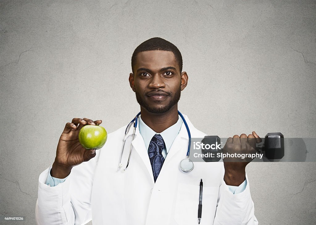 Happy doctor holding green apple, dumbbell Closeup portrait happy, smiling male health care professional, doctor in lab coat holding, lifting green apple, dumbbell, promoting, advising on healthy life style, isolated grey, black background Adult Stock Photo