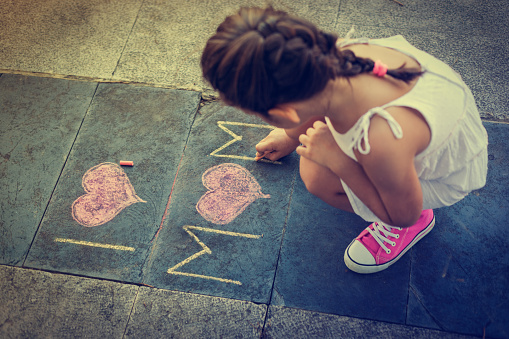 Beautiful little girl with brown hair writes on the sidewalk I love my mum, with chalk, symbolizing Mother's Day. She is wearing a white dress.Little girl is squatting and looking down while writing.