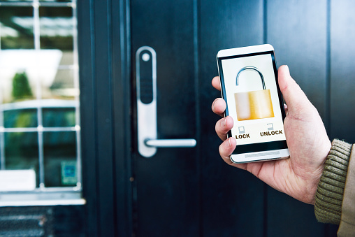 Photo of a hand holding a mobile phone. The phone shows a running app with the icon of an open lock. In the background is a front door to a house. Concept of home automation and internet of things, where products like locks connect to the internet and can be controlled remotely. Note: the image on the screen is my work.