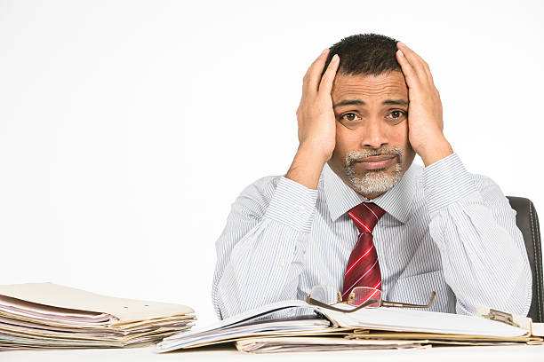 Mature businessman holding his head with his hands while a look of exasperation shows on his face. He feel hopeless and tired because of failure in business. Loads of files and paper are spread on his desk. He is in formal wear with red tie. The image is shot on white background.