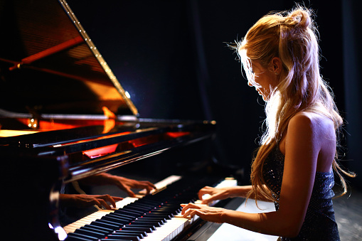 Side view of middle aged caucasian woman playing baby grand piano in a concert.She has long blond hair partially pulled back with a clip and shiny sleeveless dress.