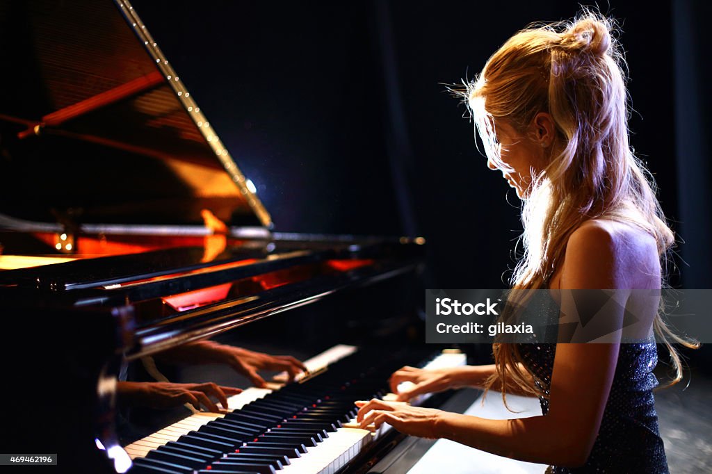 Woman playing piano in a concert. Side view of middle aged caucasian woman playing baby grand piano in a concert.She has long blond hair partially pulled back with a clip and shiny sleeveless dress. Piano Stock Photo