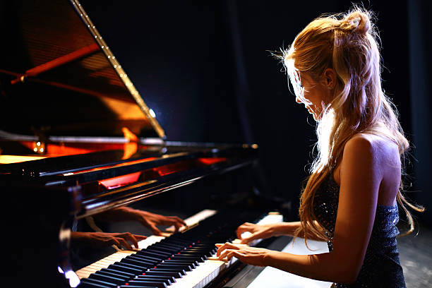 Woman playing piano in a concert. Side view of middle aged caucasian woman playing baby grand piano in a concert.She has long blond hair partially pulled back with a clip and shiny sleeveless dress. pianist stock pictures, royalty-free photos & images