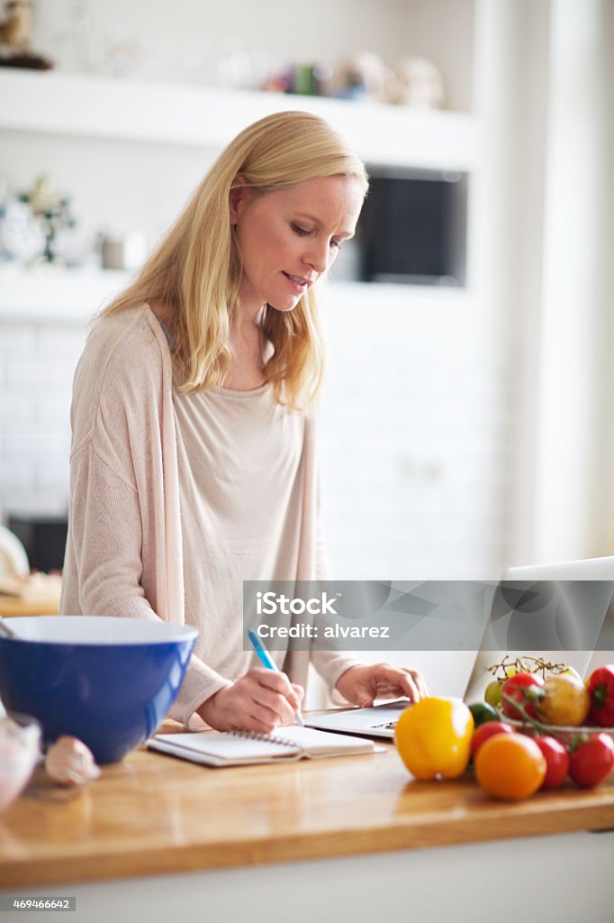 Beautiful young woman taking recipe from internet Beautiful young woman writing down recipe in a book from internet using laptop in kitchen Diary Stock Photo
