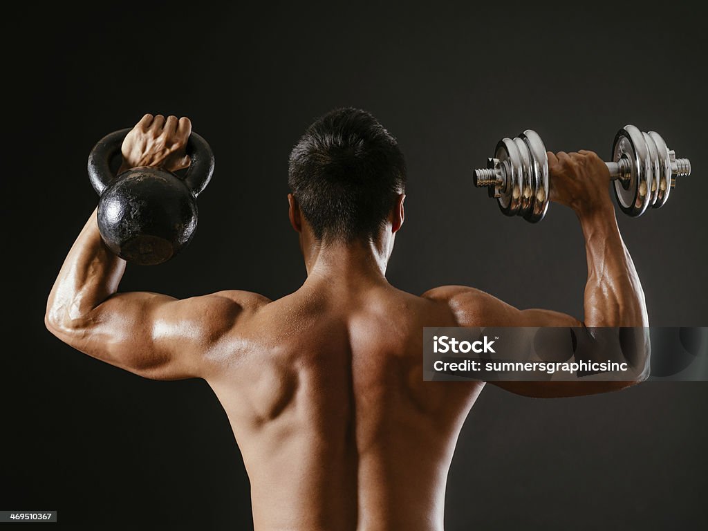 Kettlebell or dumbbell Photo of an Asian male exercising with both a kettlebell and a dumbbell, doing shoulder press over dark background. Shoulder Press Stock Photo