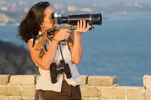 Adult female photographer shooting sea birds with an analogic film  camera equiped with telephoto mounted in a sniper rig.  The woman wears a skirt short and hunting boots resembling the safari style of clothing. The location is a high view point that looks to the coastline bays and cliffs. Photos were taken in the Pacific coast of Mexico at Huatulco, Oaxaca, She carries binoculars and sunglasses.