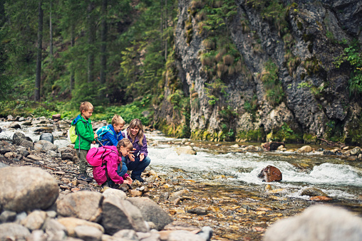 Mother and three kids hiking. The family is at the moutain river bank and there are forest and rocks in the background. Everybody is wearing jackets and backpacks. There are three kids: a girl aged 8 and two twin boys aged 5.