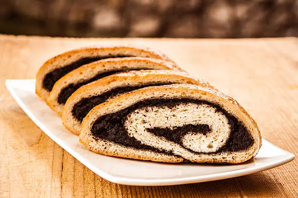 Portion of sliced fresh baked poppy seed roll served on white plate and rustic wooden table. This cake is traditional for Christmas and Easter holidays in East and South-East Europe.