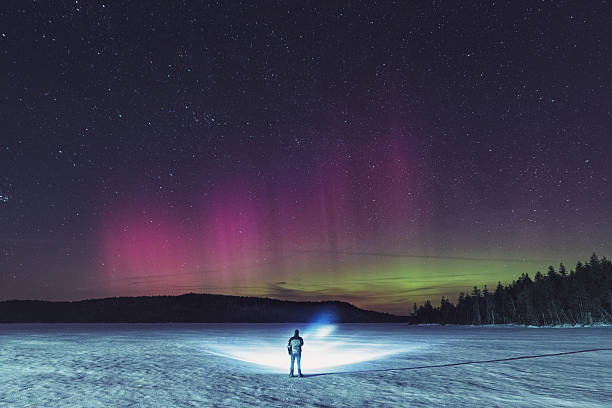Searching The North A man stands on a frozen lake searching the distant shoreline with a bright spotlight while the Northern Lights shimmer above.  Long exposure. snowshoeing snow shoe red stock pictures, royalty-free photos & images