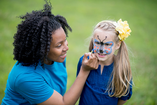 A little girl getting her face painted like a butterfly, outside at the park on a nice summer day.