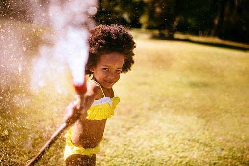 Smiling little african american girl spraying water straight at the camera from a garden hose in her back yard