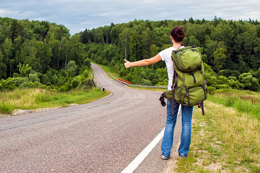 Woman with green backpack hitchhiking on a country road. Back view