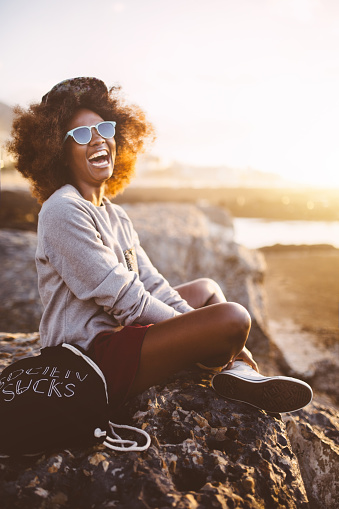 Happy Hipster Afro teen girl sitting on some rocks  on the beach at sunset and laughing