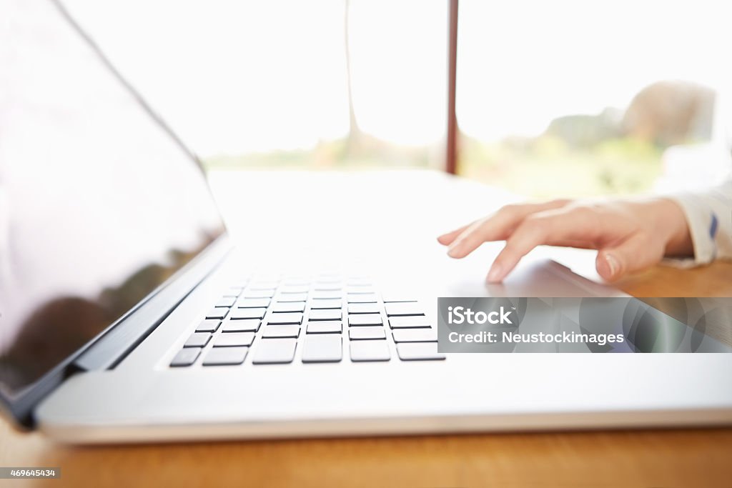 Cropped image of hand using laptop at table Cropped image of hand using laptop at wooden table. Woman is in brightly lit home. The table is found by glass window. The focus is on computer keys 2015 Stock Photo