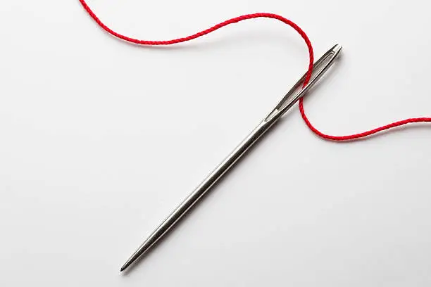A close up of the eye of a needle being thread with red thread.  The thread gracefully curves across the image as it makes its way in and out of the eye of the needle.  It is photographed on a white background
