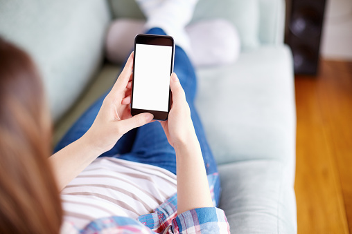 Over the shoulder view cropped image of relaxed casually dressed woman using smart phone. She is relaxing on sofa at home. The focus is on the phone with blank screen. This empty space can be used for advertisement purpose.