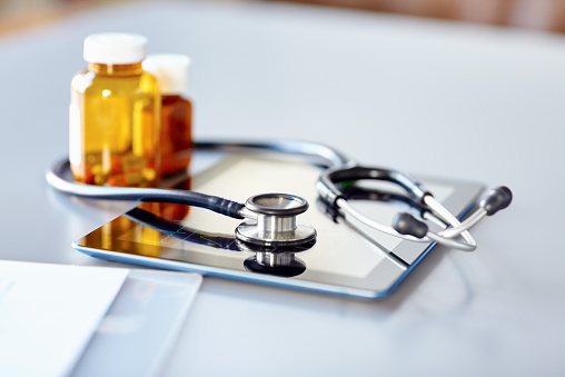 Close-up image of stethoscope with perscription pill bottle and digital tablet on desk. Focus is on chestpiece on tablet PC. Desk is in clinic.