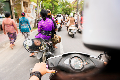 This over the shoulder royalty free, stock photograph is from a motorbike rear passenger point of view. An 18 year old Indonesian woman is driving the vehicle. Her helmet is out of focus in the foreground providing copy space. The road ahead in Ubud, Bali is full of other motor scooters and pedestrians wearing colorful, traditional Balinese clothing during late afternoon rush hour. Photographed with a Nikon D800 DSLR camera.