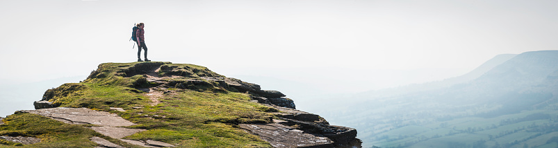 Young girl hiker standing on rocky summit looking out over a panoramic vista to the hazy mountain escarpment of the Brecon Beacons National Park, Wales, UK. ProPhoto RGB profile for maximum color fidelity and gamut.