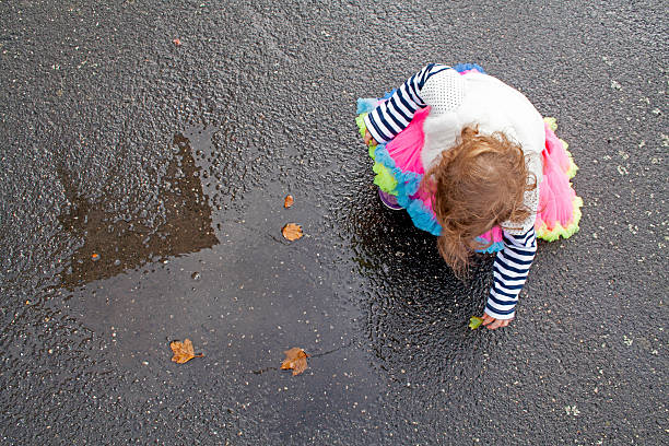 A top-down view (birdseye) of a toddler (girl) wearing brightly coloured clothing as she plays with dry Autumn Oak leaves in a fresh rain puddle on a city pavement (sidewalk) in Ballarat, Victoria (Australia).
