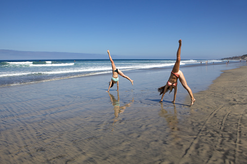 Teenage girls doing cartwheels at the beach in the wet sand.  There is a lot of copy space available in the sand and blue sky.  This was photographed in San Diego in Southern California