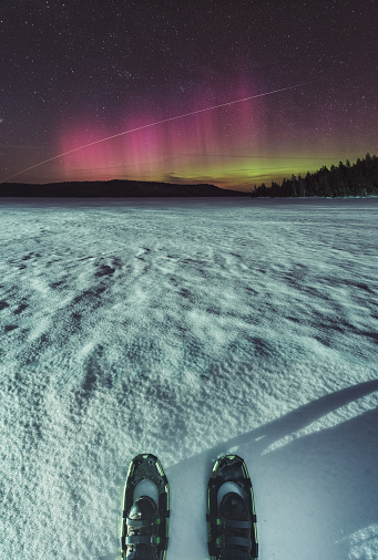 A man stands on a frozen lake wearing a pair of snowshoes watching the International Space Station streak through Northern skies painted in Aurora Borealis.  Long exposure.