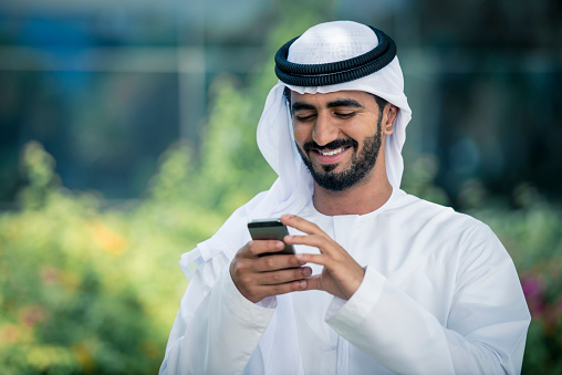 Arab businessman using mobile phone outdoots in nature. He is looking at the phone and smiling. In back, there are some green plants.