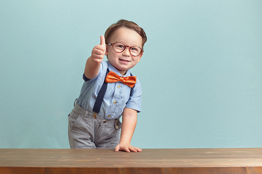 Around three years old boy in an orange bow tie and glasses, wearing blue shirt. He is smiling while giving you thumbs up over baby-blue backgorund, behind the brown table, left hand up on the table. Photo was taken by Canon DSLR.