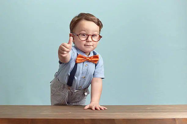 Around three years old boy in an orange bow tie and glasses, wearing blue shirt. He is smiling while giving you thumbs up over baby-blue backgorund, behind the brown table, left hand up on the table. Photo was taken by Canon DSLR.
