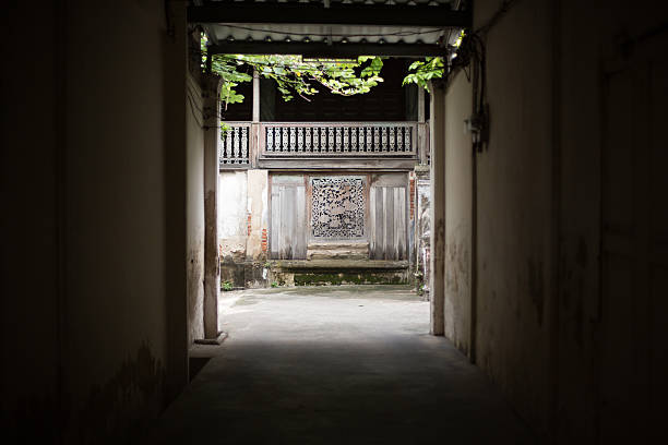 A DSLR image of the view through a dark alleyway into a brightly-lit courtyard with a wooden fench & trees, Talat Phlu, Bangkok, Thailand.