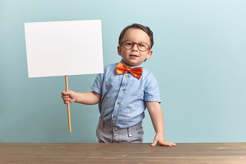 Around three years old boy in an orange bow tie and glasses, wearing blue shirt. He stands behind the brown table over baby-blue backgorund. He is holding a white banner on his right hand. He looks like happy, joyfull and confident. Photo was taken by Canon DSLR.
