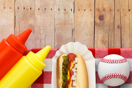 Traditional ballpark hotdog with mustard message, ketchup, onions, relish in bun on a weathered wood picnic table.  Served on a sanitary white paper fluted hot dog tray  with baseball to complete the theme.