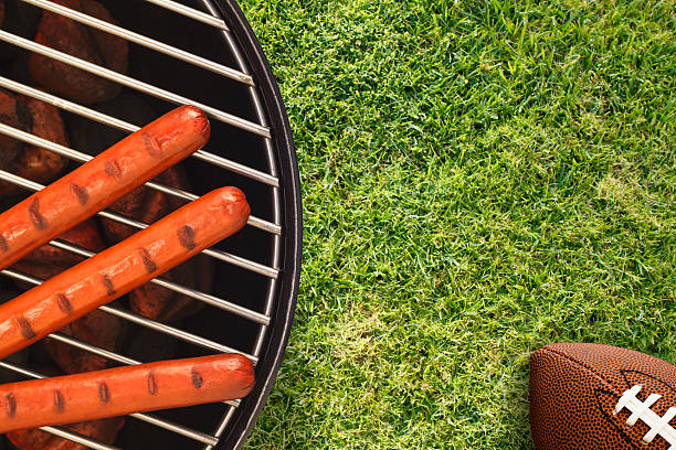 Tailgate BBQ with Hotdogs and American Football Grilling three hotdogs on a portable grill with glowing charcoal briquettes and American football in the corner.  The freshly mowed natural green turf provides copy space for the tailgate party theme tailgate party photos stock pictures, royalty-free photos & images