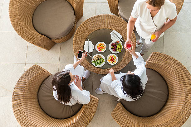 iStockalypse Dubai.  Two Arabian girlfriends in white bathrobes and slippers eating a healthy spa lunch at a resort hotel while a therapist serves them fresh orange and watermelon juice.  Aerial, overhead view.  Dubai, UAE.