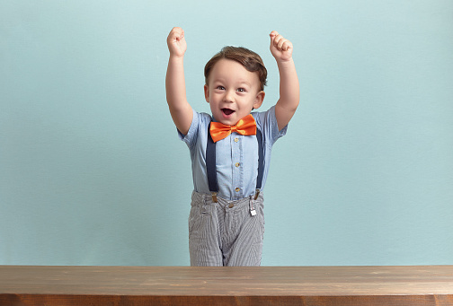 Around three years old boy in an orange bow tie and blue shirt. He put his hands up behind the brown table over baby-blue backgorund. He seems to be happy, successful and excited. Photo was taken by Canon DSLR