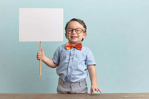 A happy little boy holding a blank sign wearing a bow tie Around three years old boy in an orange bow tie and glasses, wearing blue shirt. He stands behind the brown table over baby-blue backgorund. He is holding a white banner on his right hand. He looks like happy, joyfull and confident. Photo was taken by Canon DSLR. bow tie photos stock pictures, royalty-free photos & images