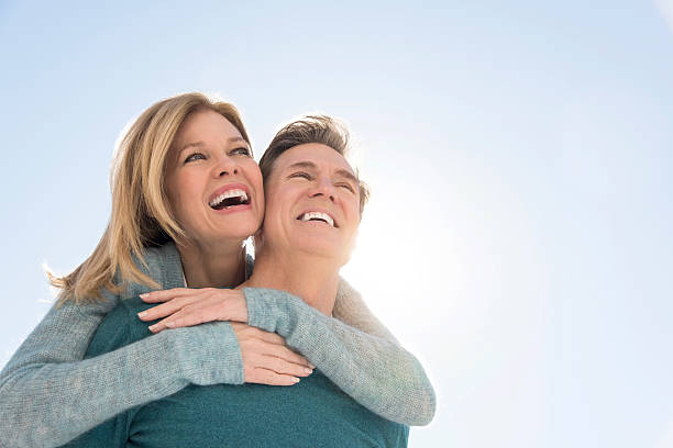 An image of a man giving a piggyback ride to a woman Happy couple against clear sky. Couple are in warm clothing. View is taken from below. Loving man giving piggyback ride to woman. 50 59 years stock pictures, royalty-free photos & images