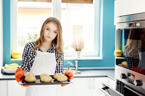 Beauty blond housewife holding hot roasting pan with hot, freshly baked bread rolls. Protecs hers hands with orange kitchen gloves