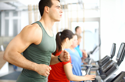 Side view of early 30's muscular caucasian guy exercising on a treadmill.He's looking straight forward. Wearing dark green sleeveless shirt.There are two women in background,blurry.