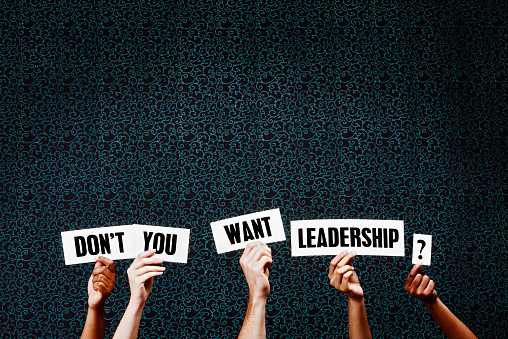 Five  hands hold up words saying 'Don't you want leadership?' against a dark background with ample copy space. Everyone needs great leadership.