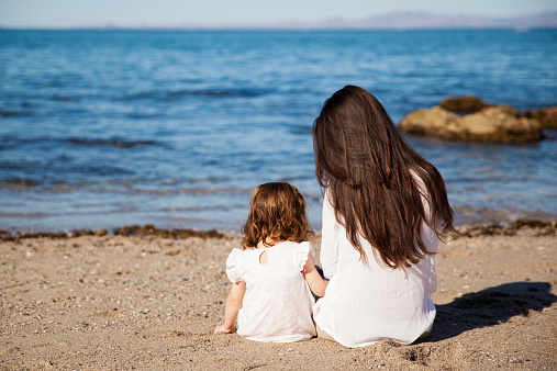 Rear view of a young brunette mom sitting next to her daughter while looking at the ocean