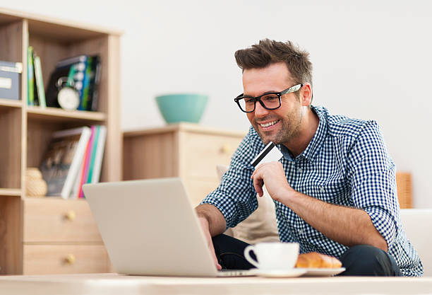 homem feliz sentado no sofá com o laptop e cartão de crédito - people joy relaxation concentration imagens e fotografias de stock