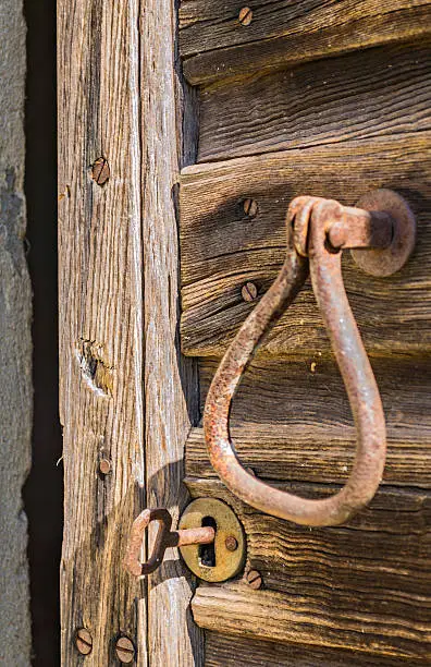 Lock of an old door belonging to an abandoned house. The door, to which is fixed a rusty doorknocker, is formed by worn and chipped wooden boards. A rusty key is inserted into the keyhole. The crumbling house is situated in the Langhe (Piedmont, Italy), UNESCO World Heritage Site.