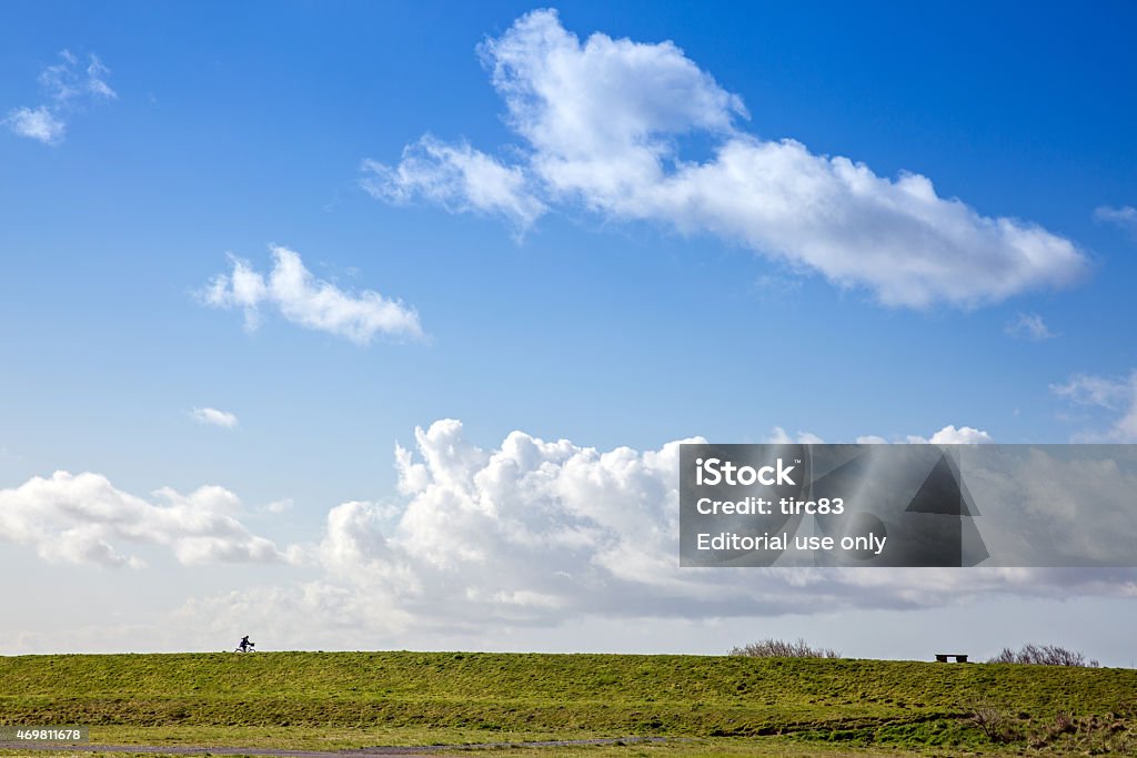 Woman cyclist with blue sky breaking through white cloud background Llanelli, UK - April 11, 2015: Woman cyclist on grassy ridge with blue sky breaking through white cloud background 2015 Stock Photo