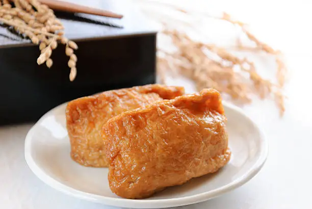 Japanese Inari-sushi, which is fried bean-curd stuffed with boiled rice. those are served on white backgound with lunch box and rice stalks,