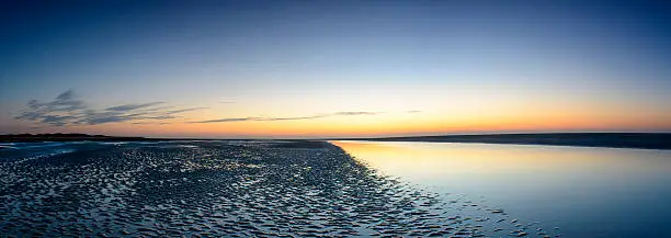 This image depicts a beach at sunset. There are very few clouds in the sky, which ranges from hues of yellow and orange to a dark blue color at the top. The colors of the sunset are reflected in the calm water which meets the dark sand in the middle of the picture.