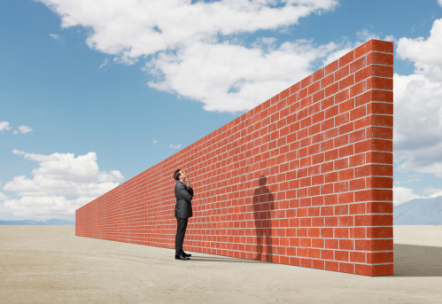 A man stands with his arms folded in front of him as he looks up towards a long and tall brick wall. The brick wall represents an impediment to his progress.  He is contemplating a solution to getting around the wall.  The wall is oddly placed in the middle of a desert salt flat .